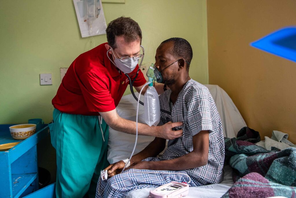 A doctor uses a stethoscope to listen to the breathing of a man wearing an oxygen mask