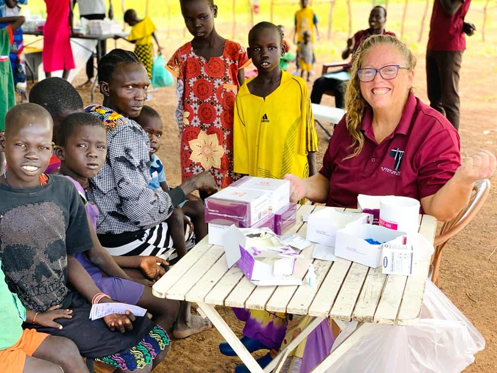 A smiling healthcare worker sitting outside with a group of Sudanese kids