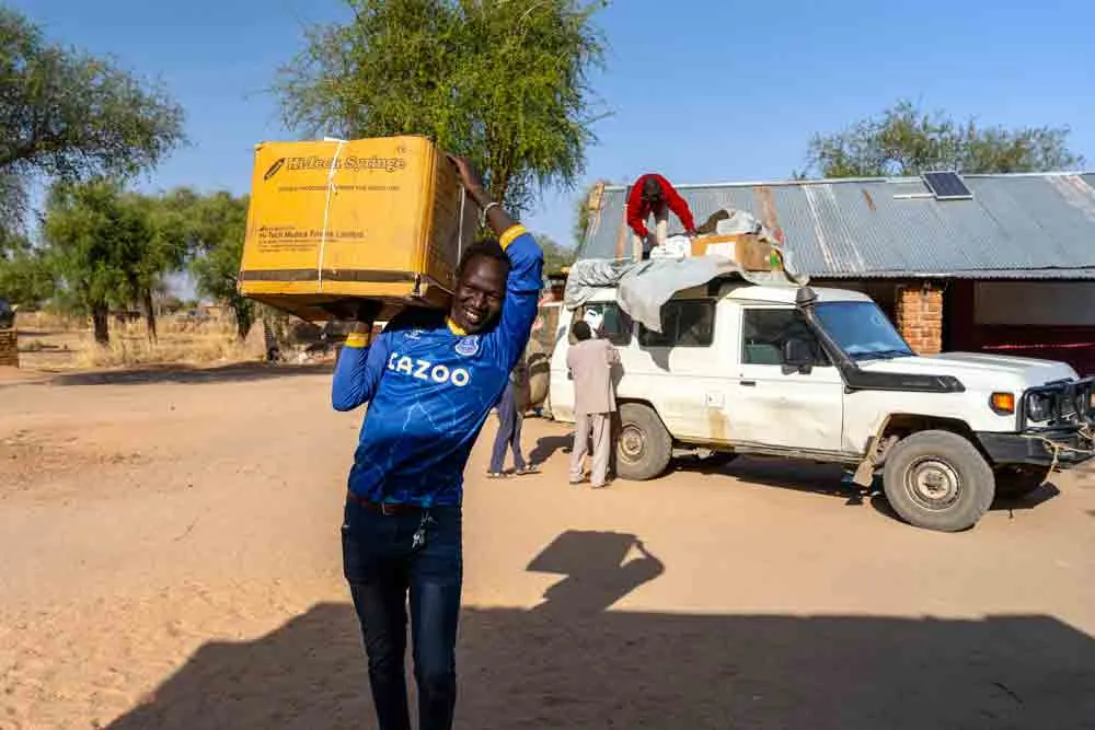A man carries a box of medical supplies near a Land Cruiser being unloaded in a rural hospital compound.